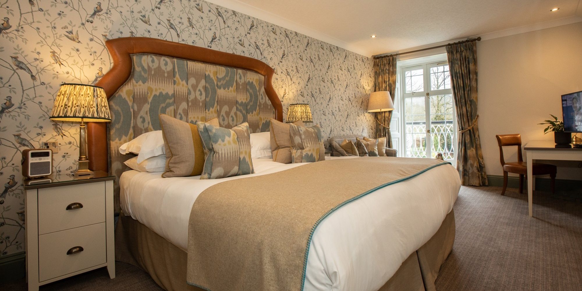 An elegant bedroom with a large bed, patterned cushions, bird-themed wallpaper and a seating area at Rothay Manor Hotel.