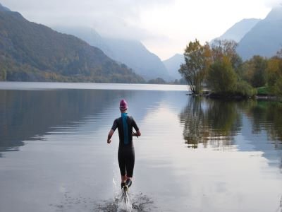 A woman in wetsuit running into a serene lake in the Lake District to enjoy wild swimming surrounded by trees and hills.